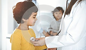 Woman-doctor examining a child patient by stethoscope in sunny clinik. Cute arab boy at physician appointment