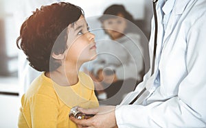 Woman-doctor examining a child patient by stethoscope in sunny clinik. Cute arab boy at physician appointment