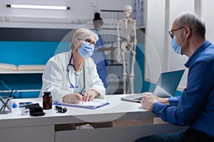 Woman doctor with face mask taking notes on papers at medical consultation with man