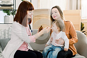 Woman doctor giving spoon dose of medicine liquid drinking syrup to child baby girl her patient. Doctor visit at home