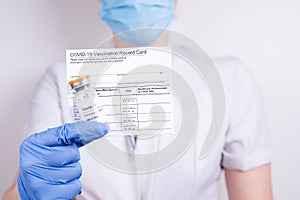A woman doctor in a mask and gloves holding a vial with covid-19 vaccine and vaccination record card