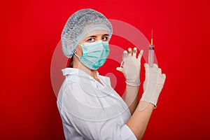 Woman doctor in medical clothes holds a syringe and an ampoule in her hands. Studio photo on a red background.