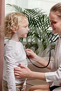 A woman doctor in a medical coat listens to small patient with a phonendoscope.