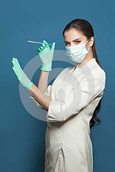 Woman doctor or nurse in professional medical uniform and protective face mask holding syringe on blue background, medicine,