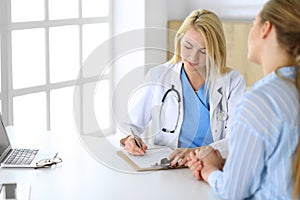 Woman doctor and patient sitting and talking at medical examination in hospital or clinic. Blonde therapist cheerfully