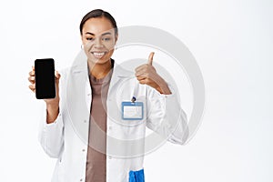 Woman doctor shows thumbs up and smartphone screen app, mobile healthcare application interface, standing over white