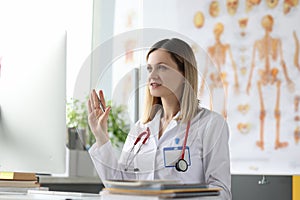 Woman doctor sit at table and waving at computer screen