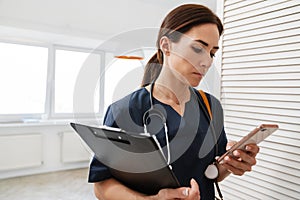 The woman doctor with stethoscope looking into the phone while holding a tablet with papers in hand