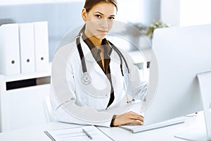 Woman-doctor typing on pc computer while sitting at the desk in hospital office. Physician at work