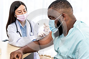 Woman doctor, wearing a surgical mask, is administering a vaccination treatment to an African American man patient