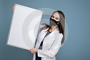 Woman doctor wearing a white medical coat and a mask holding blank board with copy space for text isolated on background
