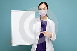 Woman doctor wearing a white medical coat and a mask holding blank board with copy space for text isolated on background