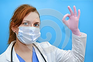 Woman doctor in white coat shows ok sign, concept on blue background. Portrait of a nurse girl, close up