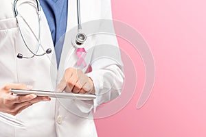 A woman doctor in a white lab coat with a pink ribbon and a stethoscope using a tablet with a pink background