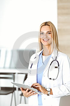 Woman-doctor at work in clinic excited and happy of her profession. Blond female physician is smiling while using tablet