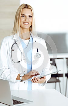 Woman-doctor at work in clinic excited and happy of her profession. Blond female physician is smiling while using tablet