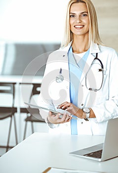 Woman-doctor at work in clinic excited and happy of her profession. Blond female physician is smiling while using tablet