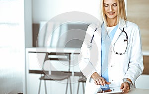 Woman-doctor at work in clinic excited and happy of her profession. Blond female physician is smiling while using tablet