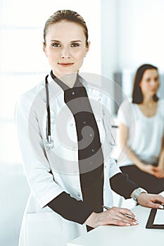 Woman-doctor at work with colleague and patient at background. Middle aged female physician filling up medical documents