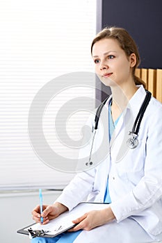 Woman doctor at work at hospital. Young female physician write prescription or filling up medical form while sitting in