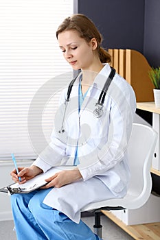 Woman doctor at work at hospital. Young female physician write prescription or filling up medical form while sitting in