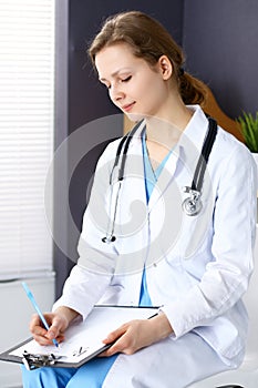 Woman doctor at work at hospital. Young female physician write prescription or filling up medical form while sitting in