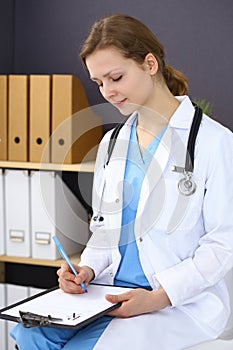 Woman doctor at work at hospital. Young female physician write prescription or filling up medical form while sitting in