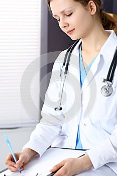 Woman doctor at work at hospital. Young female physician write prescription or filling up medical form while sitting in