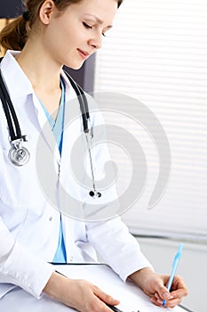 Woman doctor at work at hospital. Young female physician write prescription or filling up medical form while sitting in