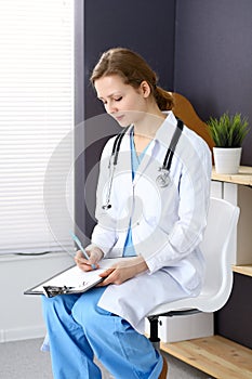 Woman doctor at work at hospital. Young female physician write prescription or filling up medical form while sitting in