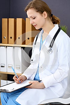 Woman doctor at work at hospital. Young female physician write prescription or filling up medical form while sitting in