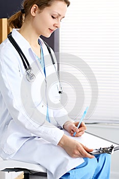 Woman doctor at work at hospital. Young female physician write prescription or filling up medical form while sitting in