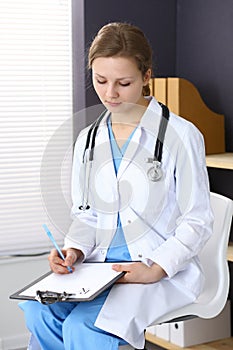 Woman doctor at work at hospital. Young female physician write prescription or filling up medical form while sitting in