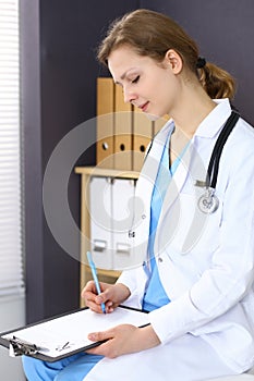 Woman doctor at work at hospital. Young female physician write prescription or filling up medical form while sitting in