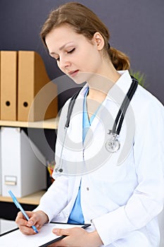 Woman doctor at work at hospital. Young female physician write prescription or filling up medical form while sitting in