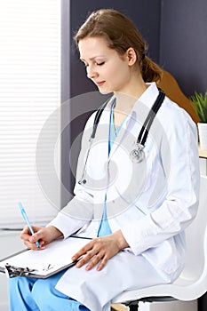 Woman doctor at work at hospital. Young female physician write prescription or filling up medical form while sitting in