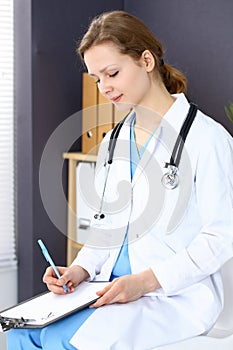 Woman doctor at work at hospital. Young female physician write prescription or filling up medical form while sitting in