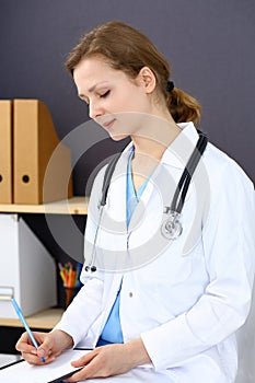 Woman doctor at work at hospital. Young female physician write prescription or filling up medical form while sitting in