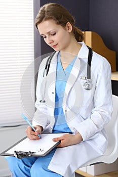 Woman doctor at work at hospital. Young female physician write prescription or filling up medical form while sitting in