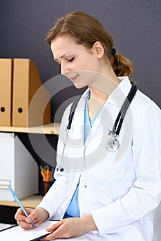 Woman doctor at work at hospital. Young female physician write prescription or filling up medical form while sitting in