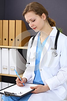 Woman doctor at work at hospital. Young female physician write prescription or filling up medical form while sitting in