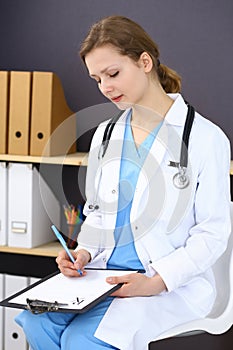 Woman doctor at work at hospital. Young female physician write prescription or filling up medical form while sitting in