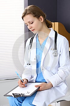 Woman doctor at work at hospital. Young female physician write prescription or filling up medical form while sitting in