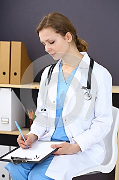 Woman doctor at work at hospital. Young female physician write prescription or filling up medical form while sitting in