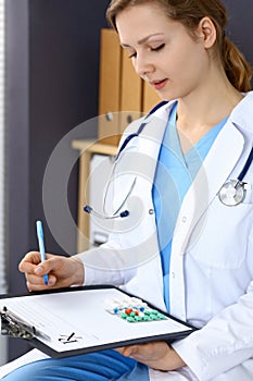 Woman doctor write prescription while sitting in hospital office. A lot of various colored pills liye at her clipboard