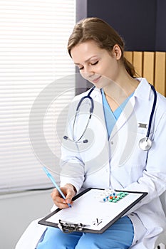 Woman doctor write prescription while sitting in hospital office. A lot of various colored pills liye at her clipboard