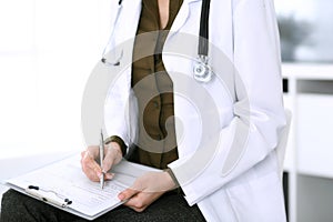 Woman doctor writing something at clipboard while sitting at the chair, close-up. Therapist at work filling up