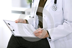 Woman doctor writing something at clipboard while sitting at the chair, close-up. Therapist at work filling up