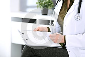 Woman doctor writing something at clipboard while sitting at the chair, close-up. Therapist at work filling up