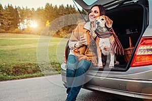 Woman and dog with shawls sits together in car trunk on autumn